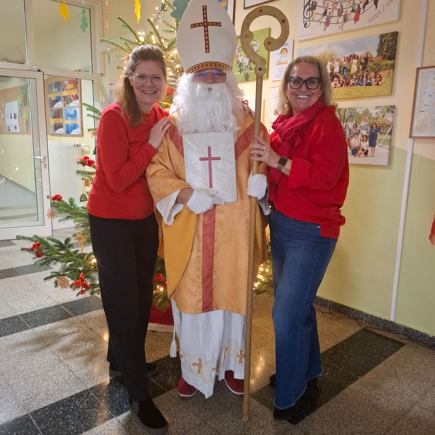 Zwei Frauen und ein Mann in einer Heiligenverkleidung stehen vor einem geschmückten Weihnachtsbaum. Sie lächeln und scheinen für ein Foto zu posieren.
