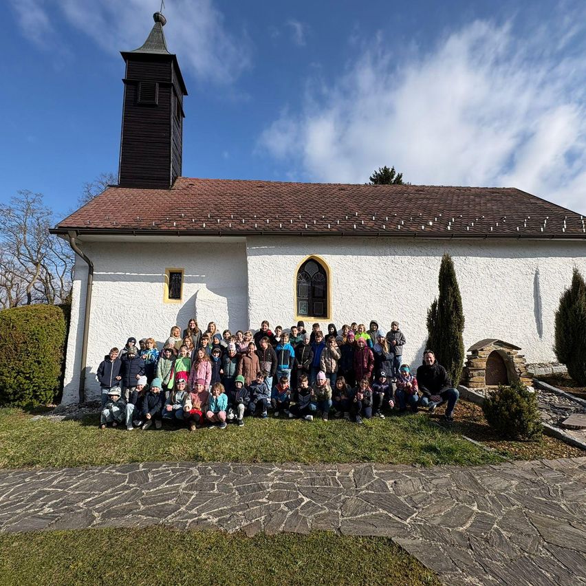 Eine Gruppe von Menschen versammelt sich vor einer weißen Kirche mit einem braunen Dach. Sie sitzen und stehen auf einem Rasen vor der Kirche.