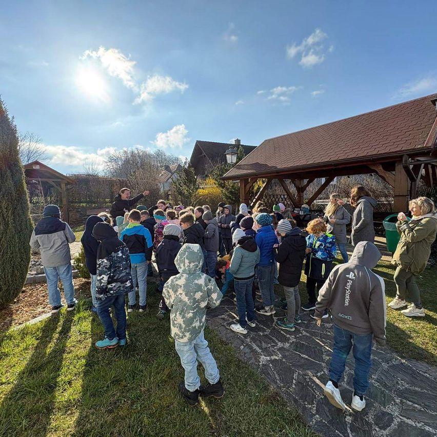 Eine Gruppe von Kindern und Erwachsenen versammelt sich unter einem sonnigen Himmel im Freien. Sie befinden sich in der Nähe eines Gebäudes mit einem braunen Dach, möglicherweise einem Gemeindezentrum.