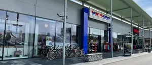 Ein Intersport-Geschäft mit einer Glaswand, die Fahrräder und eine Frau in Sportbekleidung zeigt. Eine Reihe von Fahrrädern ist vor dem Geschäft geparkt.