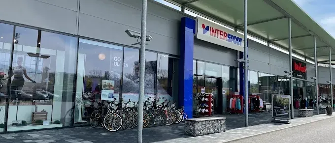 Ein Intersport-Geschäft mit einer Glaswand, die Fahrräder und eine Frau in Sportbekleidung zeigt. Eine Reihe von Fahrrädern ist vor dem Geschäft geparkt.