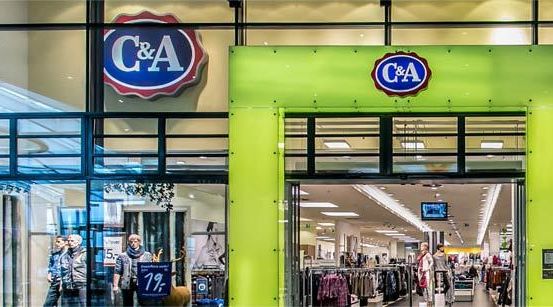 Der Eingang eines C&A-Kleidunggeschäfts zeigt eine hellgrüne Tür und Glaswände. Das Ladenschild zeigt das Logo prominent. Im Inneren hängen Kleidungsstücke an Kleiderbügeln und Mannequins tragen Jacken. Ein Verkaufsschild weist auf 17% Rabatt hin.