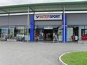 Der Eingangsbereich eines Intersport-Geschäfts. Der Außenbereich zeigt ein großes Schild, eine blau-weiße Struktur und einen Rasenbereich. In der Nähe sind Fahrräder geparkt.