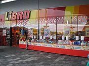Die Vorderseite eines Ladens namens LIBRO ist leuchtend bunt in Rot und Gelb. Im Inneren sind Regale mit Büchern und anderen Artikeln gefüllt. Der Laden hat ein großes Fenster, das die Umgebung widerspiegelt.
