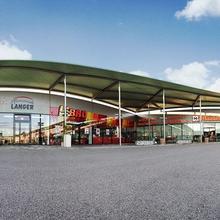 Ein Freiluft-Einkaufszentrum mit einem grünen Vordach, mit Geschäften wie Langer, BBQ und Miller. Das Gebiet ist von einer Rasenfläche und einer leeren Asphaltfläche umgeben.