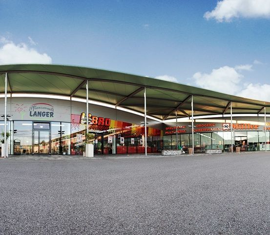 Ein Freiluft-Einkaufszentrum mit einem grünen Vordach, mit Geschäften wie Langer, BBQ und Miller. Das Gebiet ist von einer Rasenfläche und einer leeren Asphaltfläche umgeben.