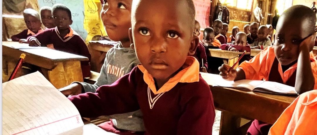 Ein Klassenzimmer mit Kindern, die an hölzernen Schreibtischen sitzen, ein junger Junge in einem Weinroten Hemd schaut in die Kamera und hält ein Notizbuch und einen Bleistift.