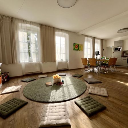 Ein geräumiges Wohnzimmer mit Holzboden, rundem grünem Teppich und Kissen drumherum. Der Essbereich ist mit Tisch und Stühlen ausgestattet. Die Küche ist mit Geräten ausgestattet. Der Raum hat Fenster mit Vorhängen.