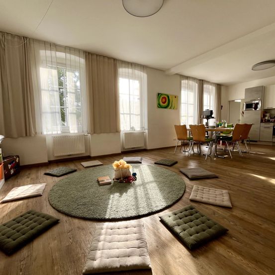 Ein geräumiges Wohnzimmer mit Holzboden, rundem grünem Teppich und Kissen drumherum. Der Essbereich ist mit Tisch und Stühlen ausgestattet. Die Küche ist mit Geräten ausgestattet. Der Raum hat Fenster mit Vorhängen.