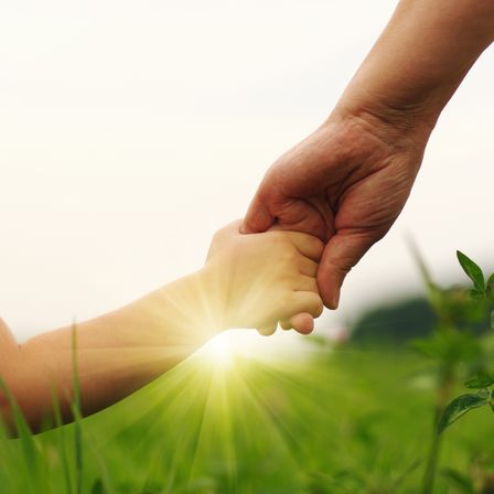 Eine Hand eines Erwachsenen hält die Hand eines Kindes auf einem Grasfeld unter Sonnenlicht.