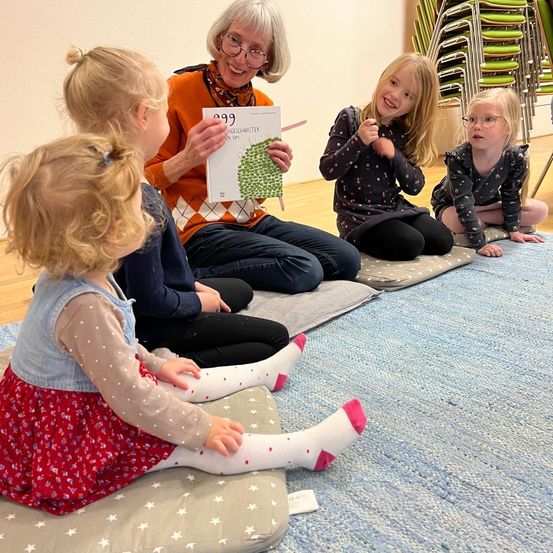 Eine Gruppe junger Mädchen sitzt auf dem Boden und hört einer älteren Frau zu, die ein Buch liest. Jedes Mädchen sitzt auf einem Kissen. Eines der Mädchen trägt ein rotes Kleid.