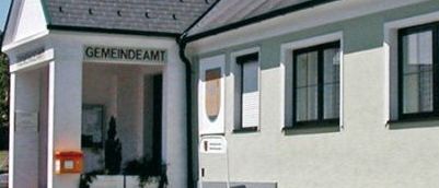 Das Äußere eines weißen Gebäudes mit grauem Dach und schwarzen Fensterrahmen, beschriftet mit 'Gemeindeamt'. Es hat drei Fenster rechts und zwei links, mit einem Baum im Hintergrund.