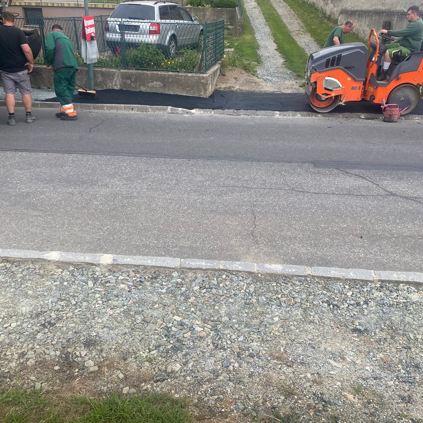 Arbeiter versiegeln eine Straße mit einem Straßenkehrer. Eine Person bedient den Kehrer, während andere die Straßenseite reinigen. Ein Auto ist in der Nähe geparkt.