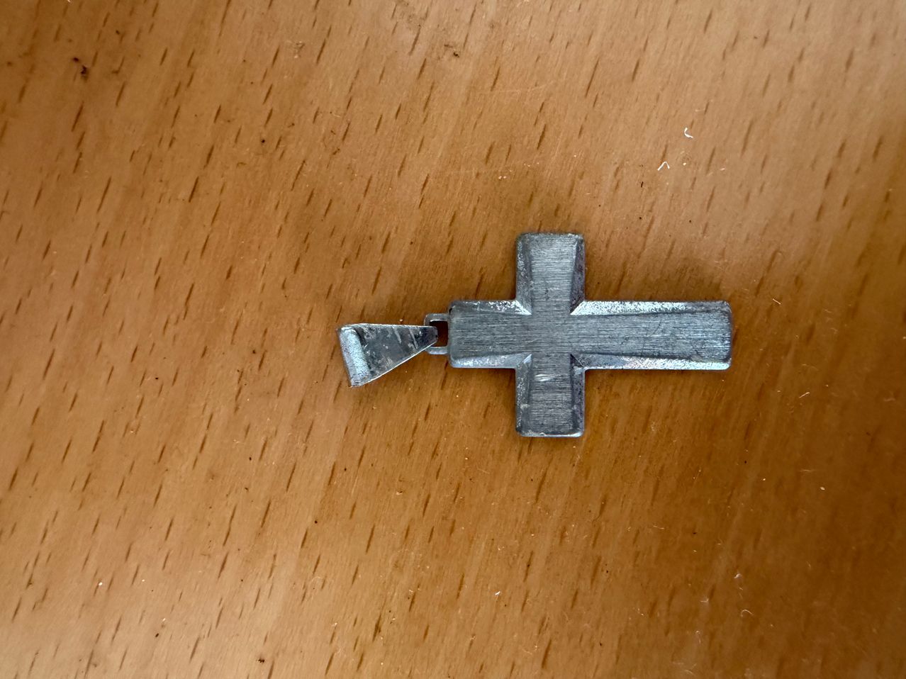 Ein kleiner, silberner Kreuzanhänger mit einer Schlaufe auf der Rückseite liegt auf einem Holztisch.
