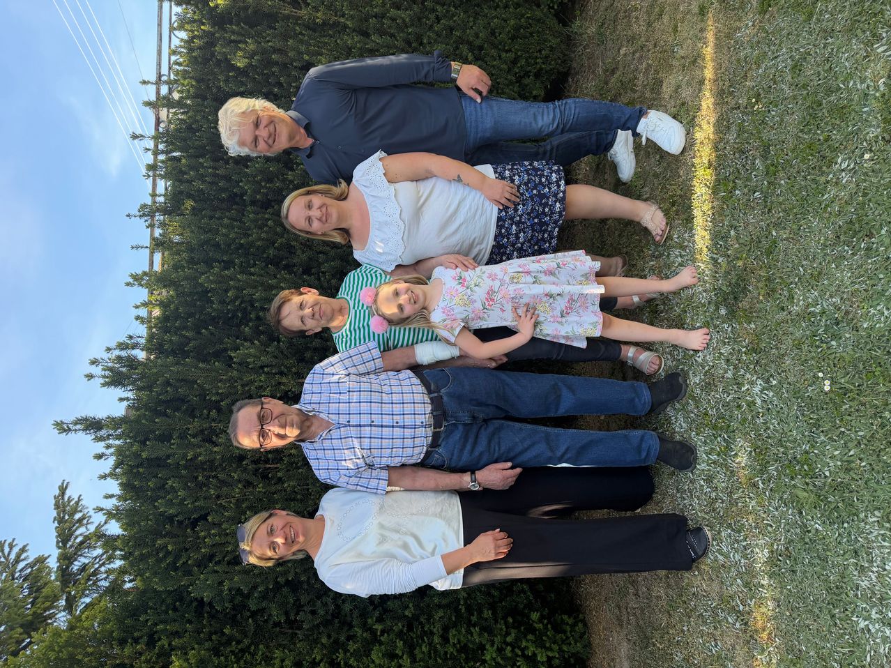 Eine Familie posiert für ein Foto in einem Garten mit grünen Büschen und einem klaren Himmel im Hintergrund. Ein Mann und eine Frau sitzen auf dem Boden, während zwei Kinder vor ihnen stehen.