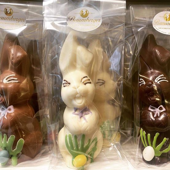 Vier Schokoladen-Osterhasen sind in Plastikverpackungen ausgestellt. Die Hasen sind mit Schokolade überzogen und haben Osterdekorationen wie Eier und Pflanzen. Die Verpackung hat ein Etikett mit dem Wort Baumberger.
