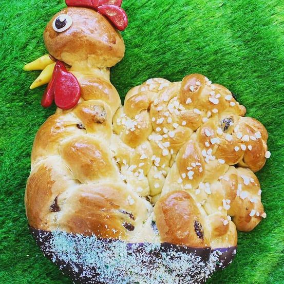 Eine geformte Brotskulptur in Form eines Huhns mit roten Kämmen und mit Sesam bestreut liegt auf einer grünen Oberfläche.