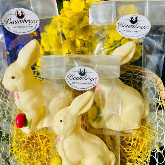Drei cremefarbene Osterhasen in Plastikverpackung sitzen in einem mit Heu gefüllten Korb. Jeder Hase hat ein kleines rotes Objekt auf dem Kopf. Hinter den Hasen sind gelbe Blumen.