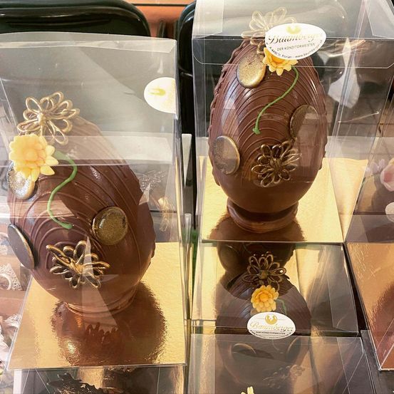 Zwei Schokoladen-Oster Eier mit dekorativen Blumen und Münzen sind in Glas-Fällen. Ein Ei hat gelbe Blumen, und das andere hat goldene Münzen. Sie sitzen auf goldenen Basen.