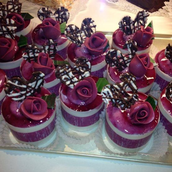 Mehrere Cupcakes mit roter Glasur und Schokoladen-Akzenten, jeweils gekrönt von einer dekorativen violetten Rose. Sie sind auf einem silbernen Tablett über einer weißen Oberfläche angeordnet.