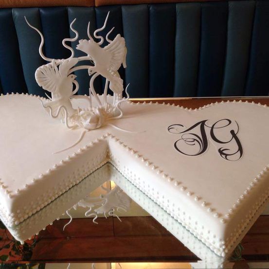 Ein herzförmiger Kuchen mit komplizierten weißen Zuckerguss-Designs, einschließlich Vögeln, ist auf einer Glasfläche ausgestellt. Er hat Perlen um den Rand und 'JG' in kursiver Schrift auf der Oberseite geschrieben.