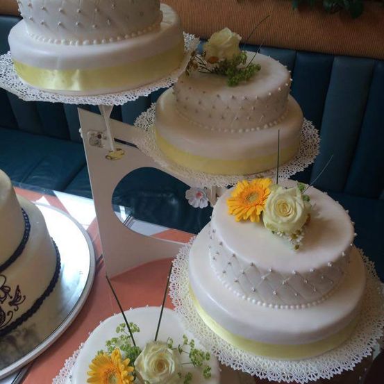 Drei weiße und gelbe Kuchen mit weißer Glasur und Blumendekorationen sind auf einem Kuchenständer ausgestellt. Die Kuchen haben komplizierte Designs und Perlenakzente.