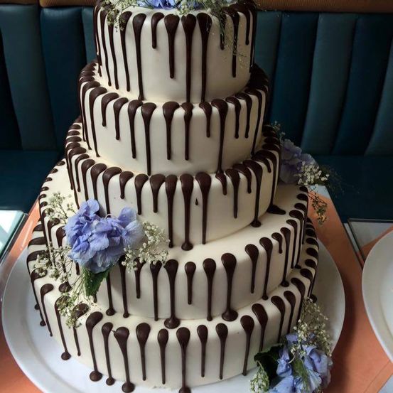 Ein dreistöckiger Kuchen mit Schokoladentropfen und blauen Blumen auf jeder Etage, auf einem weißen Teller mit blauer Polsterung im Hintergrund.