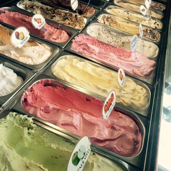 Eine Auslage mit verschiedenen Eissorten in Behältern, darunter Mango und Apfel, mit beschrifteten Löffeln.