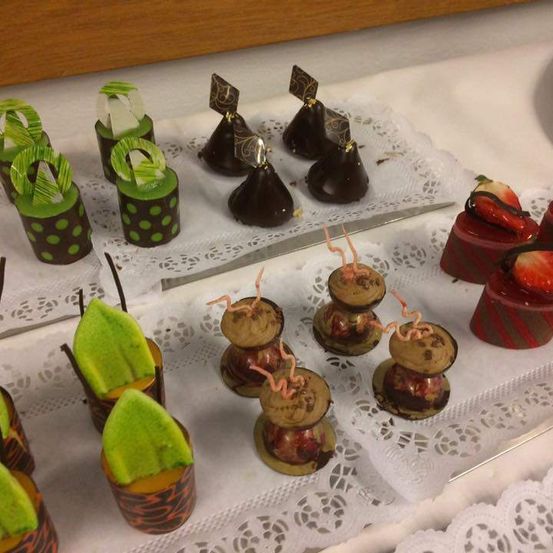 Verschiedene Desserts sind auf einem Tisch mit weißer Spitze angeordnet. Es gibt Schokoladen, grüne und rote Leckereien und eine Erdbeere. Die Schokoladen haben rote und grüne Toppings.