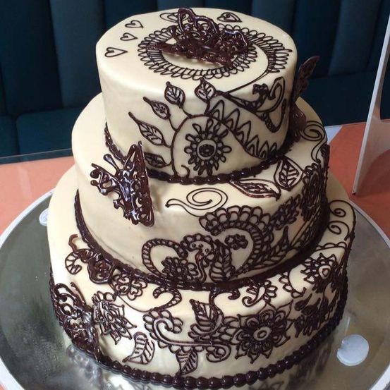 Ein dreistufiger Kuchen mit kunstvollen Schokoladenglasur-Designs von Blumen und Schmetterlingen auf einem silbernen Teller.