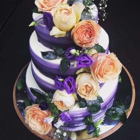 Ein dreistufiger Kuchen mit violetten und weißen Schichten, dekoriert mit gelben, orangefarbenen und violetten Rosen und grünen Blättern. Er steht auf einer silbernen Platte.