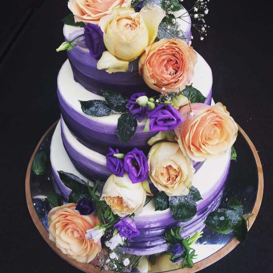 Ein dreistufiger Kuchen mit violetten und weißen Schichten, dekoriert mit gelben, orangefarbenen und violetten Rosen und grünen Blättern. Er steht auf einer silbernen Platte.