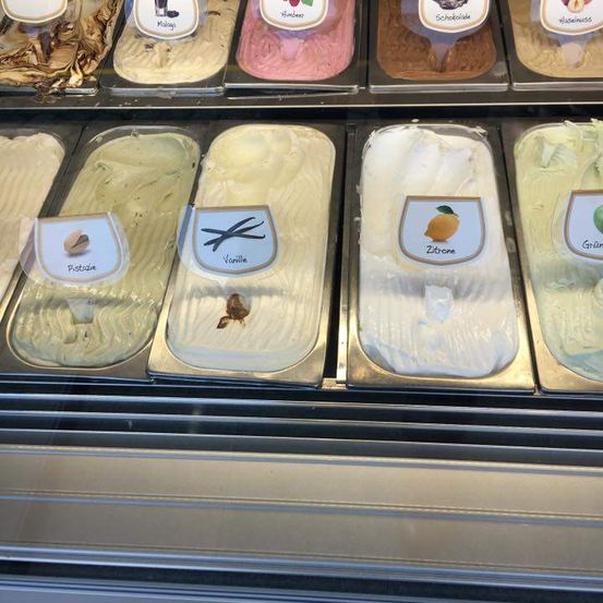 Eine Eiscreme-Auslage mit verschiedenen Geschmacksrichtungen, darunter Pistazie, Vanille und Zitrone, jeweils in eigenen Fächern.
