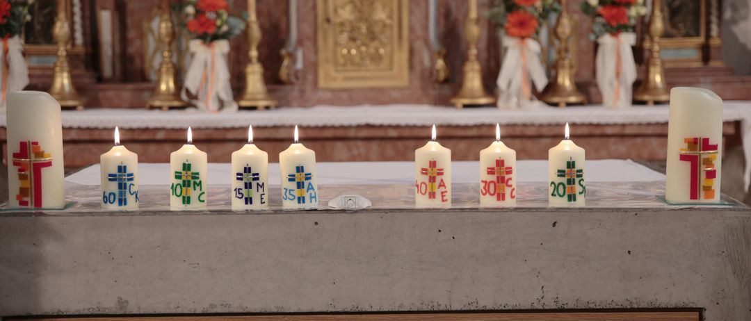 Eine Reihe von Kerzen mit verschiedenen Designs steht auf einem Holztisch vor einem Kirchenaltar, der mit Blumen geschmückt ist.
