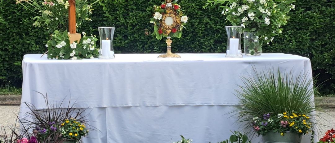 Ein mit weißem Tuch und Blumen bedeckter Tisch steht in einem Garten. Ein Kreuz und ein goldenes Objekt stehen auf dem Tisch. Zwei Kerzenhalter mit Kerzen befinden sich auf beiden Seiten.