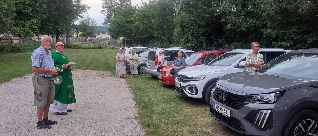 Eine religiöse Zeremonie mit geparkten Autos auf einem Rasen. Ein Mann mit einer Schüssel und ein Priester mit einem Buch stehen links.