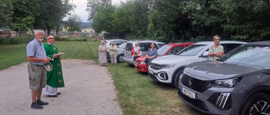 Eine religiöse Zeremonie mit geparkten Autos auf einem Rasen. Ein Mann mit einer Schüssel und ein Priester mit einem Buch stehen links.