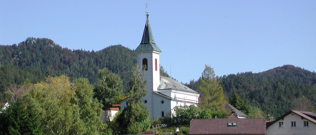 Eine malerische Dorfszene mit einer weißen Kirche und einem rotgedeckten Haus, umgeben von üppiger Grünfläche und Bergen im Hintergrund.