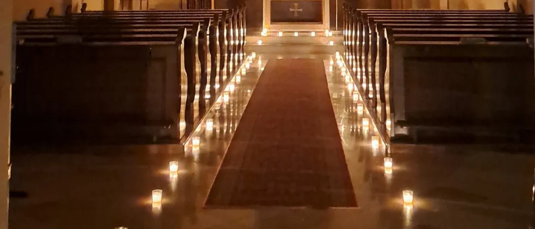 Ein schwach beleuchteter Kirchengang mit Reihen von Kirchenbänken, einem zentralen Gang mit Kerzen und einem Altar, der mit religiösen Artefakten geschmückt ist.