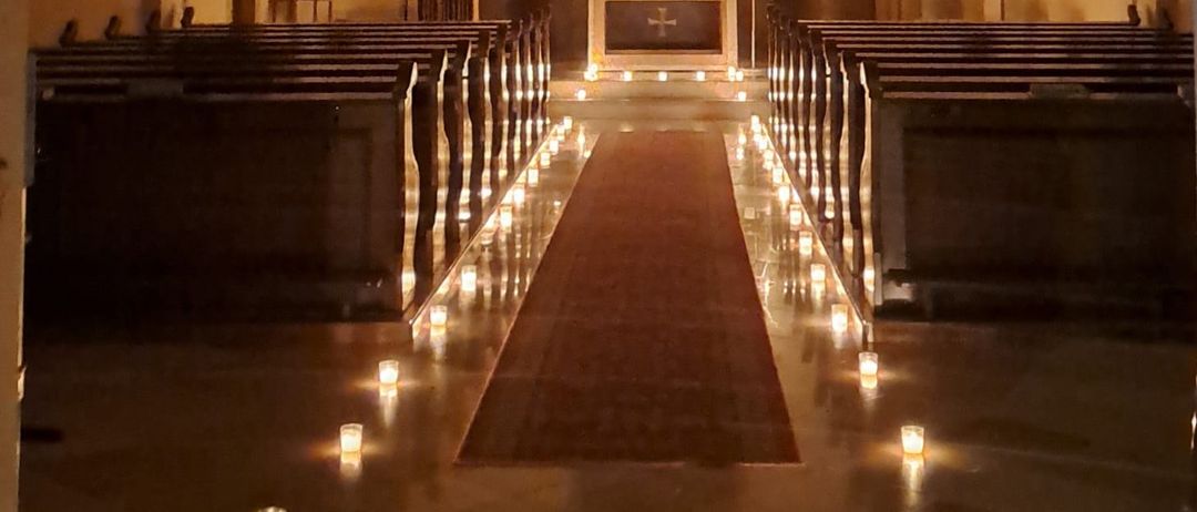 Ein schwach beleuchteter Kirchengang mit Reihen von Kirchenbänken, einem zentralen Gang mit Kerzen und einem Altar, der mit religiösen Artefakten geschmückt ist.