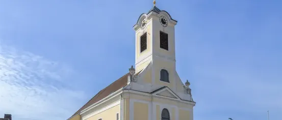 Eine Kirche mit einem spitzen Turm, einer Uhr und Rundbogenfenstern. Sie hat ein braunes Dach und ist in Gelb und Weiß gestrichen.