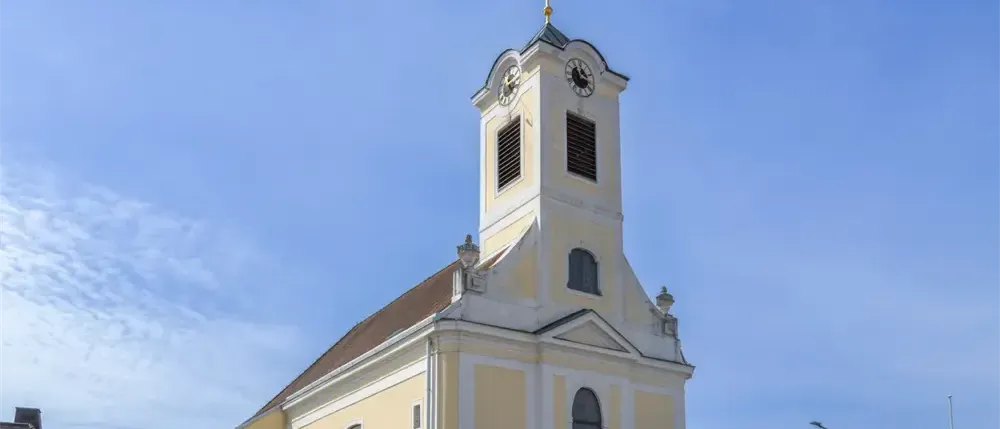 Eine Kirche mit einem spitzen Turm, einer Uhr und Rundbogenfenstern. Sie hat ein braunes Dach und ist in Gelb und Weiß gestrichen.