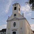 Eine weiße Kirche mit einem Glockenturm und Bogenfenstern steht an einer Straße. Ein Verkehrslicht und ein Gebäude befinden sich auf der linken Seite.