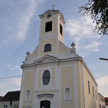 Eine weiße Kirche mit einem Glockenturm und Bogenfenstern steht an einer Straße. Ein Verkehrslicht und ein Gebäude befinden sich auf der linken Seite.