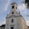 Eine weiße Kirche mit einem Glockenturm und Bogenfenstern steht an einer Straße. Ein Verkehrslicht und ein Gebäude befinden sich auf der linken Seite.