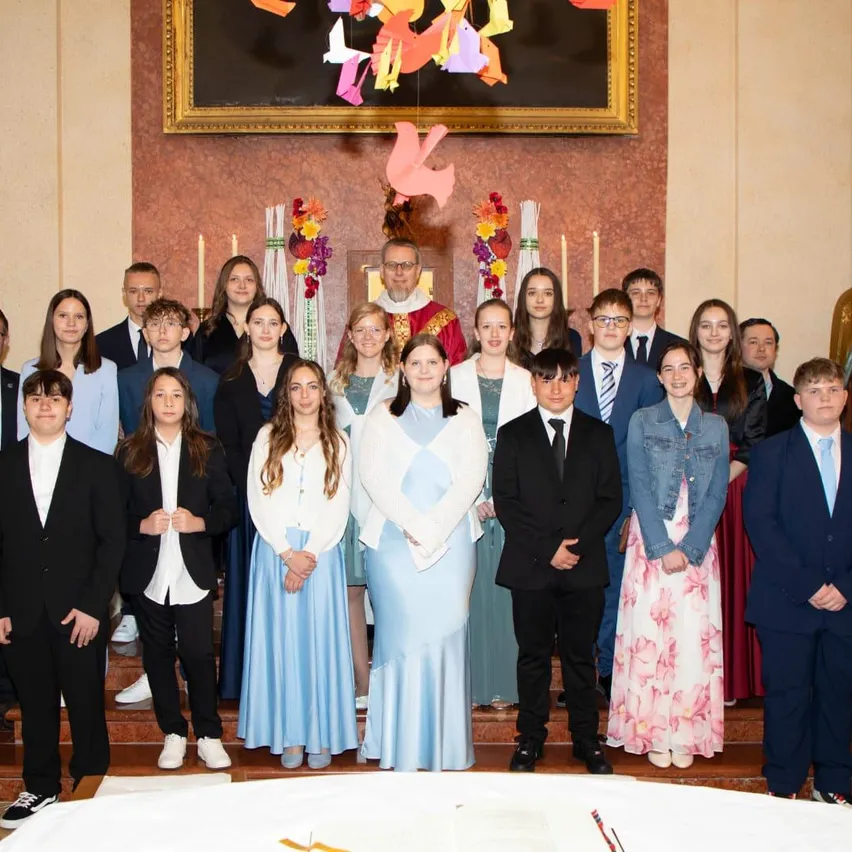 Eine Gruppe von Schülern in formeller Kleidung posiert für ein Foto vor einem Altar mit Kerzen und einem Priester in einer Kirche.
