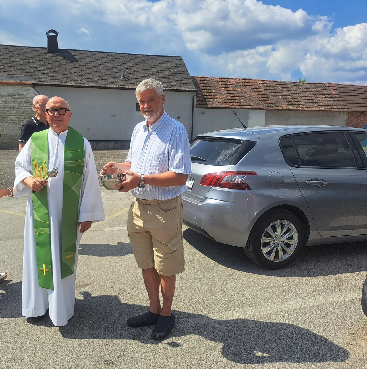 Ein Priester in einer grünen Stola steht neben einem Mann in einem gestreiften Hemd, hält eine Schüssel und im Hintergrund sind ein silbernes Auto und Gebäude zu sehen.