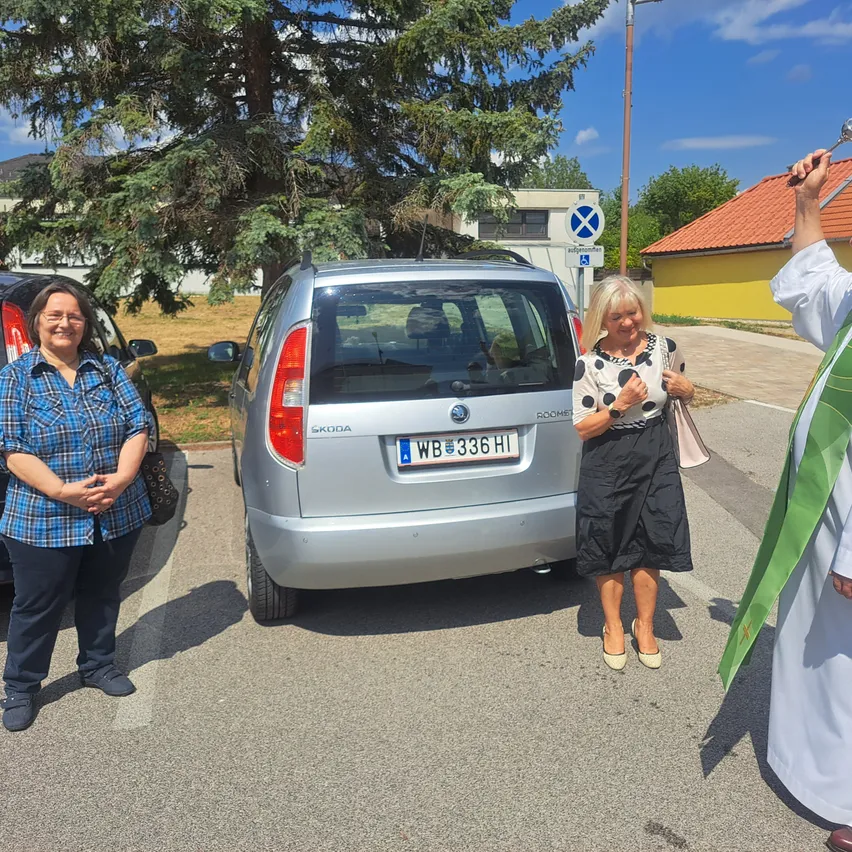 Drei Frauen stehen auf einem Parkplatz, eine hält eine Glocke, in der Nähe eines silbernen Skoda-Autos mit dem Kennzeichen 'WB 336 HI'. Ein Gebäude mit rotem Dach ist im Hintergrund zu sehen.
