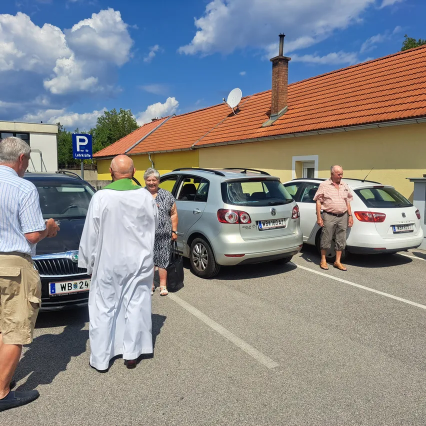 Ein Priester steht auf einem Parkplatz mit drei geparkten Autos. Der Mann im weißen Gewand spricht mit einer älteren Frau. Dahinter ist ein gelbes Gebäude mit einem roten Dach und einem Schornstein zu sehen.