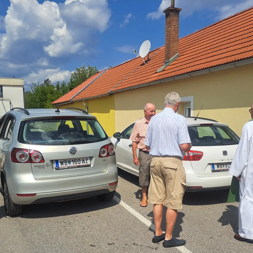 Zwei Männer stehen vor einem gelben Haus mit einem roten Dach. Ein Mann trägt ein gestreiftes Hemd und Shorts, der andere trägt ein rosa Hemd und Shorts. Ein Auto mit einem Nummernschild ist vor dem Haus geparkt.